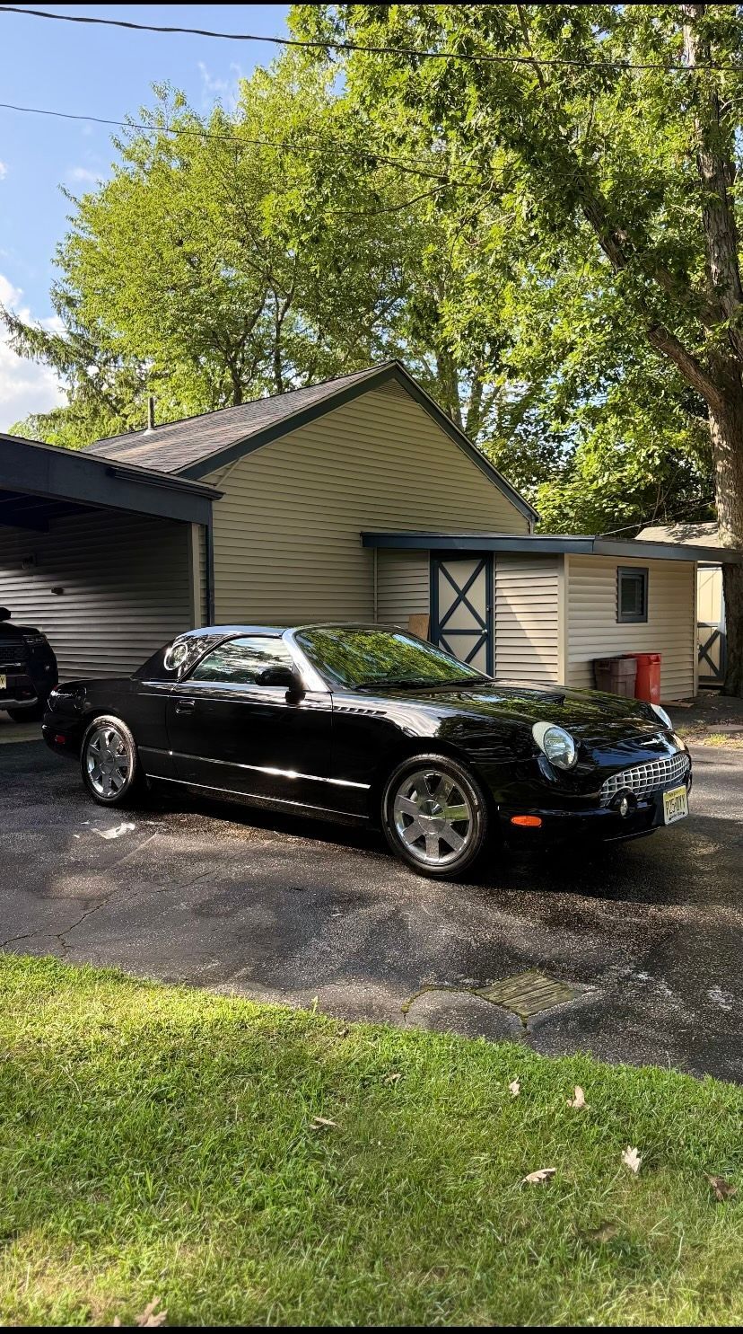 Black convertible parked in a driveway beside a tan garage under leafy trees.
