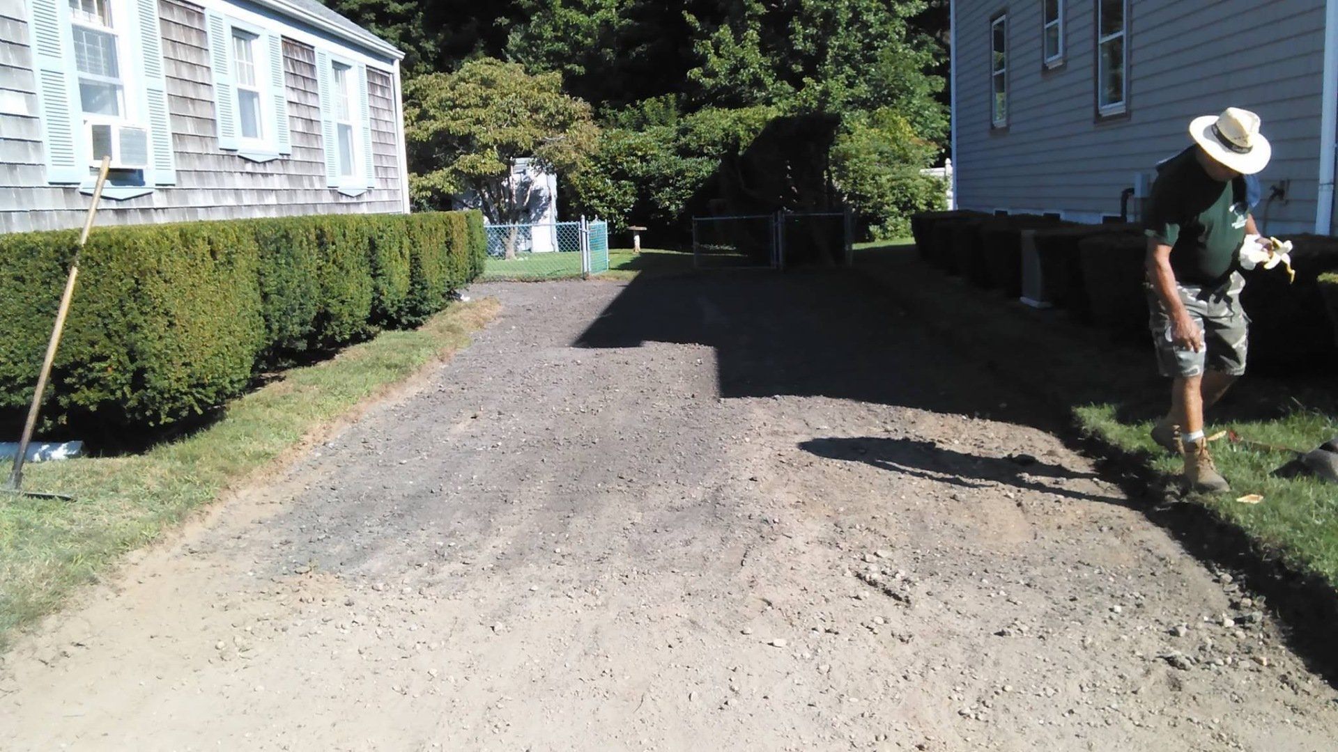 A man in a hat is standing on a dirt road in front of a house