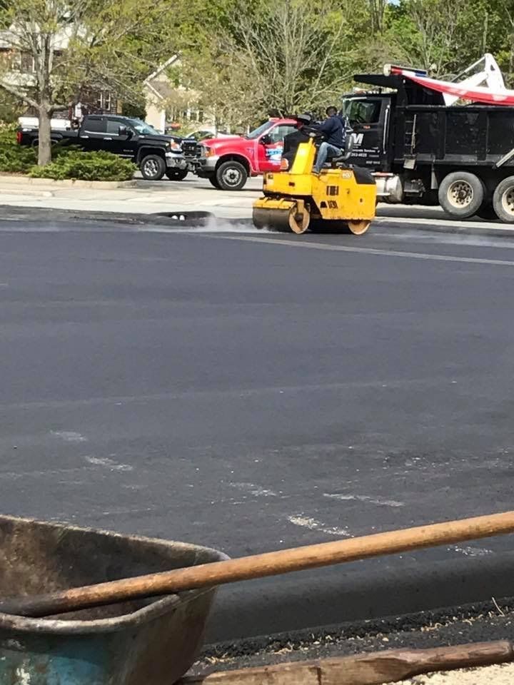 A man is driving a yellow roller on a road next to a dump truck.