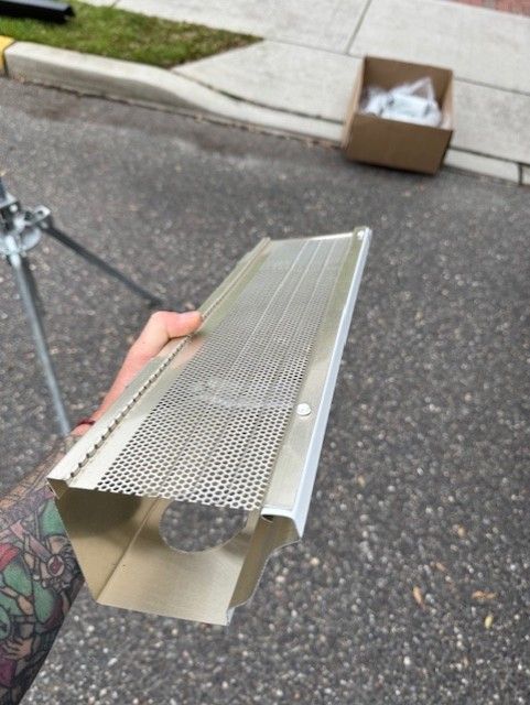 Hand holding a silver gutter guard with a perforated metal screen. Box and tripod visible.