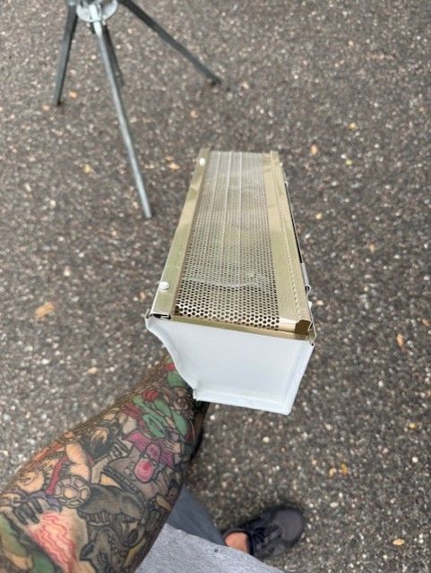 Person holding a white gutter with a gold mesh cover outdoors, a tripod visible in the background.