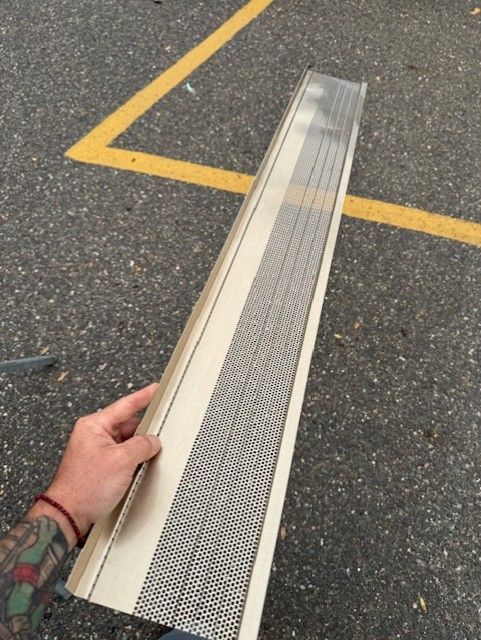 A hand holding a tan gutter guard with a perforated metal mesh section, outdoors on pavement.