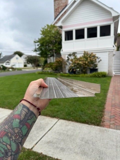 Hand holding a clear plastic soffit section outdoors. House with white siding in background.