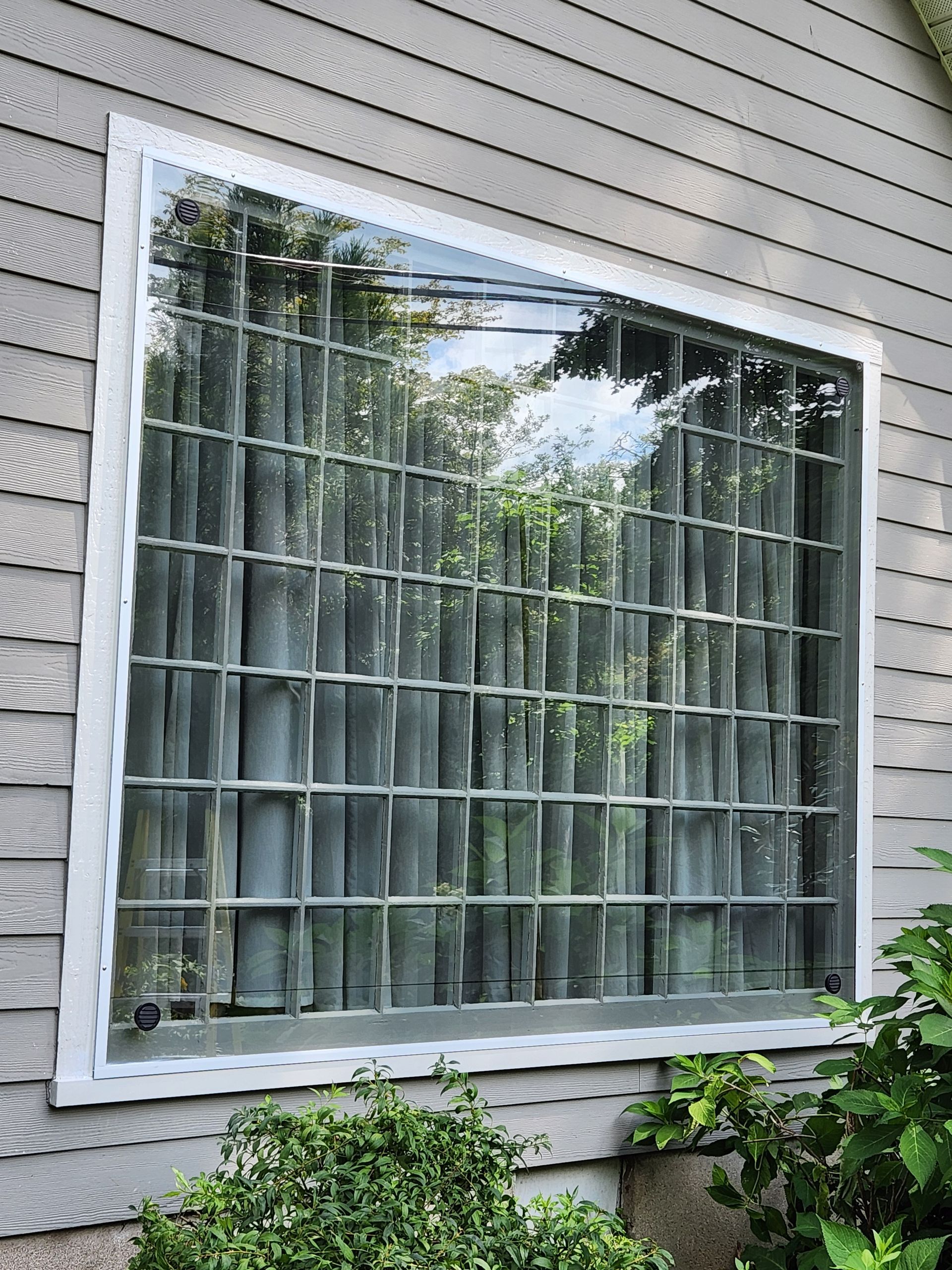 Large white-framed window with vertical blinds on a gray-sided house exterior.