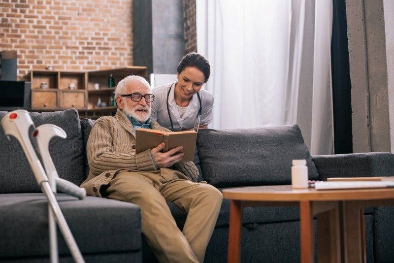 An elderly man with crutches is sitting on a couch reading a book while a nurse looks on.