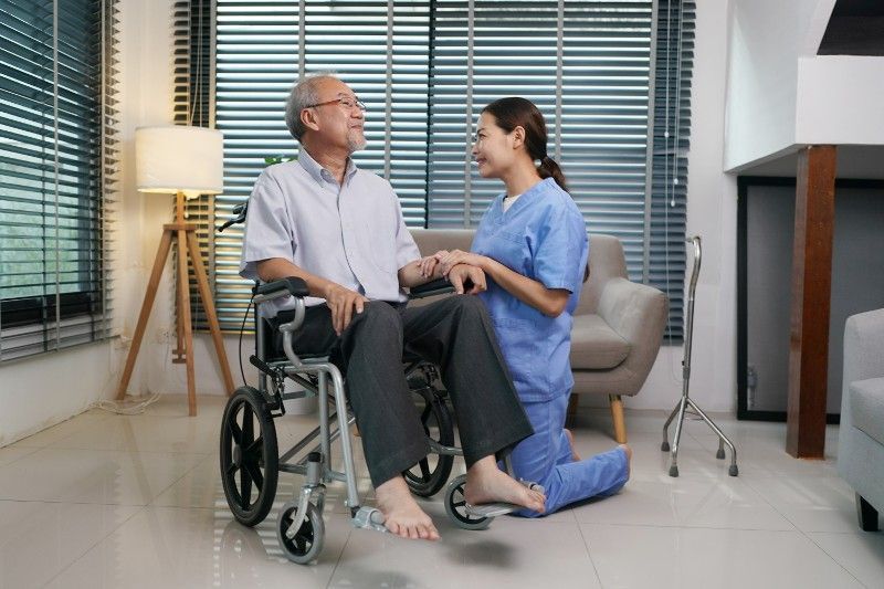 A nurse is kneeling down next to an elderly man in a wheelchair.