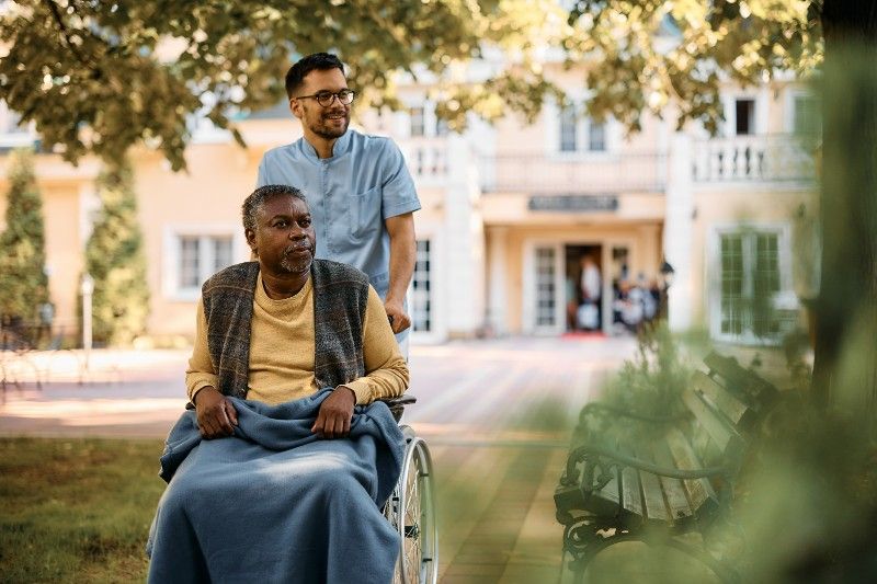 An elderly man in a wheelchair is pushed by a nurse