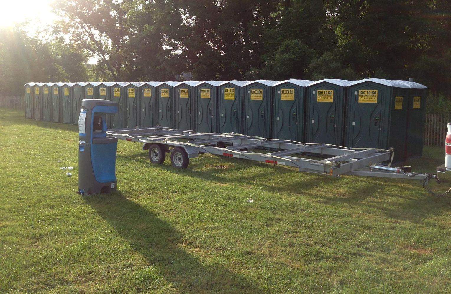 Row of green portable toilets in a grassy area, a trailer in front, with the sun shining behind.