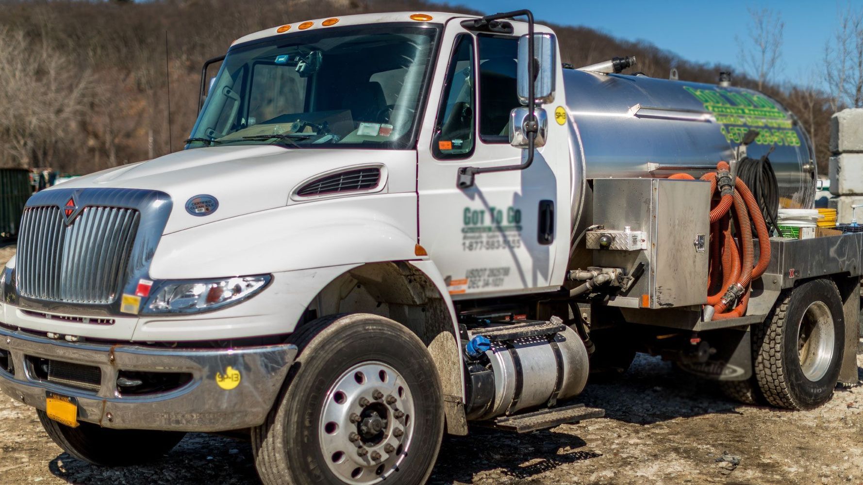 White septic truck parked outdoors on gravel. Silver tank on flatbed, orange hose.