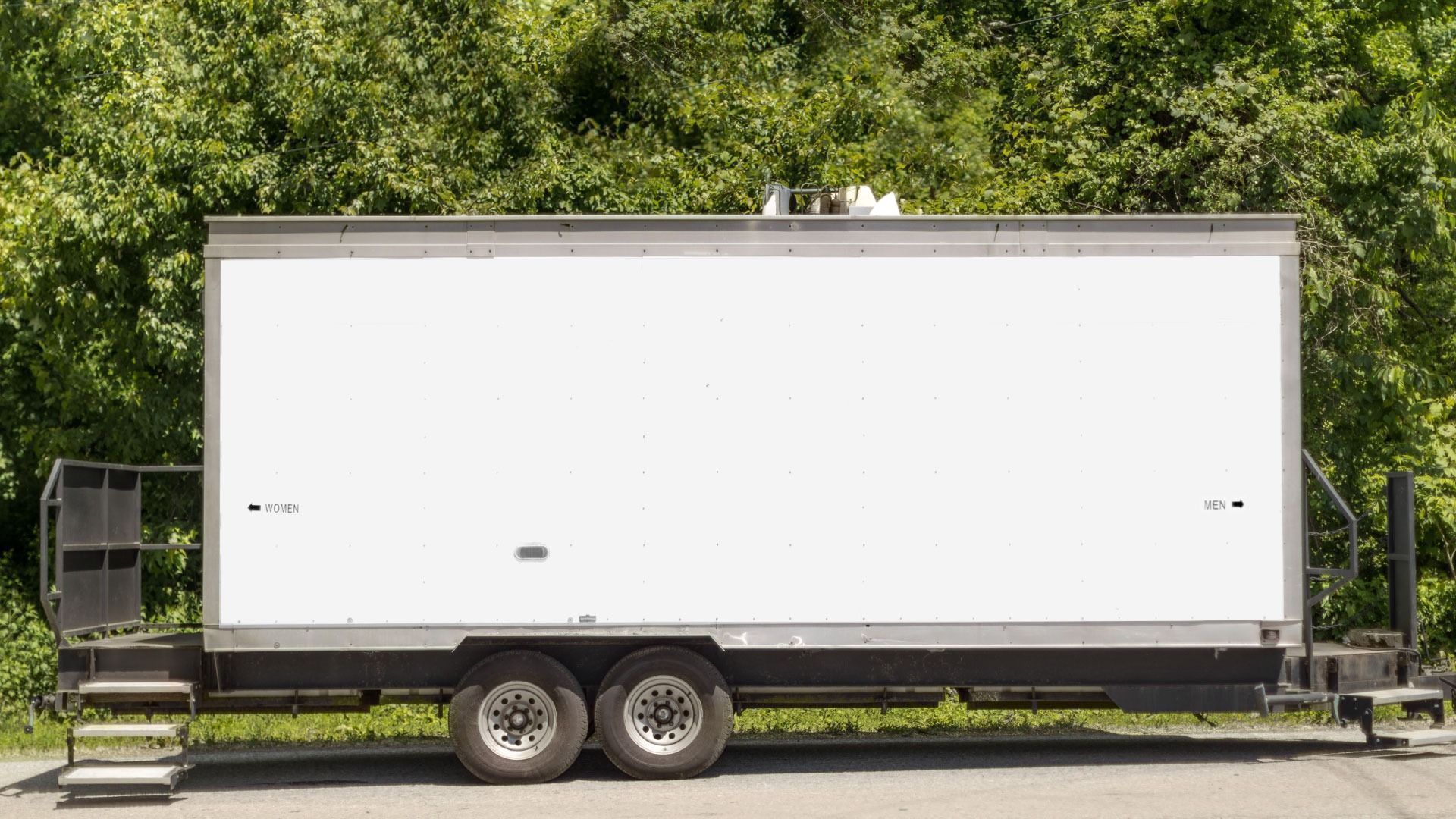White portable restroom trailer on a trailer with steps, set in front of green trees.