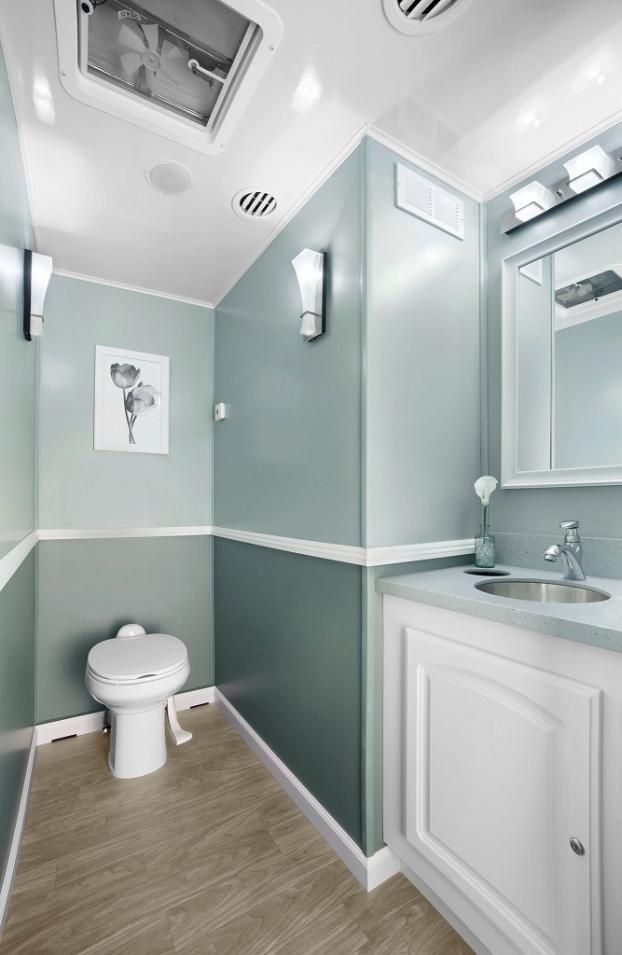 Interior of a modern portable restroom; blue-green walls, white trim, toilet, vanity, and artwork.