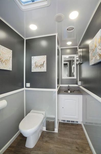 Interior of a portable restroom with gray walls, white trim, a toilet, sink, and decorative art.