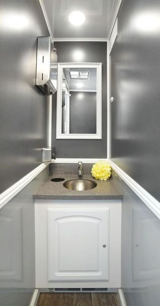 Interior of a luxury portable restroom. Gray walls, white trim, vanity with sink, mirror, soap dispenser, and paper towels.
