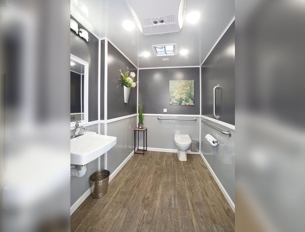 Accessible portable restroom. Gray walls, wood-look floor, sink, toilet, handrails, and flowers.