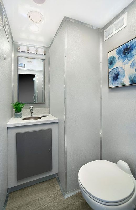 Interior of a portable restroom with a sink, mirror, toilet, and artwork. Gray walls and flooring.