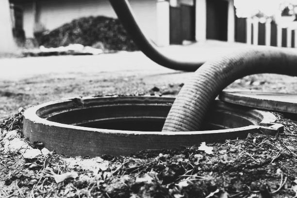 Hose entering an open manhole in a yard, debris around the opening, black and white.