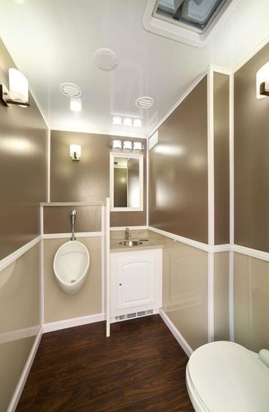 Interior of a modern portable restroom. Beige and brown walls, wood-look floor, urinal, toilet, sink, mirror.