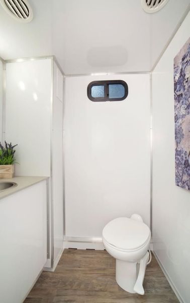 Interior of a clean, white portable restroom, featuring a toilet, sink, and small window.