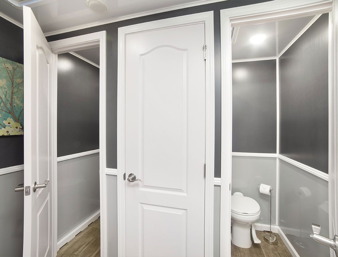 Three white-walled restroom stalls in a trailer; two doors open, one with a toilet inside.