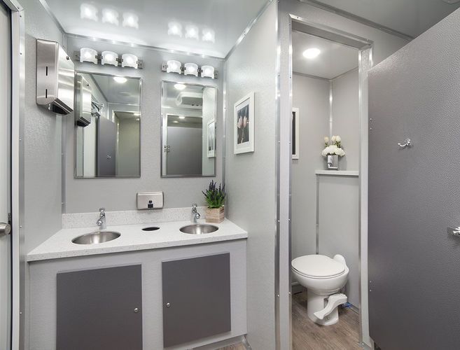 Interior of a luxury portable restroom with two sinks, mirrors, and a toilet. Light gray walls and decor.