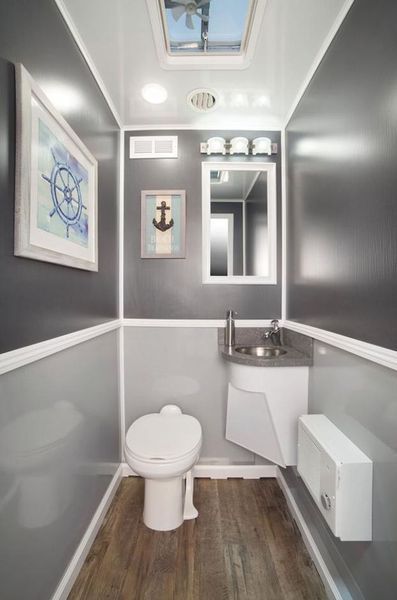 Interior of a portable restroom with nautical decor: gray walls, white trim, toilet, sink, mirror, art, and a skylight.