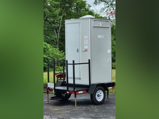 Portable toilet on a trailer; gray walls, white wheels, black frame, outdoors.