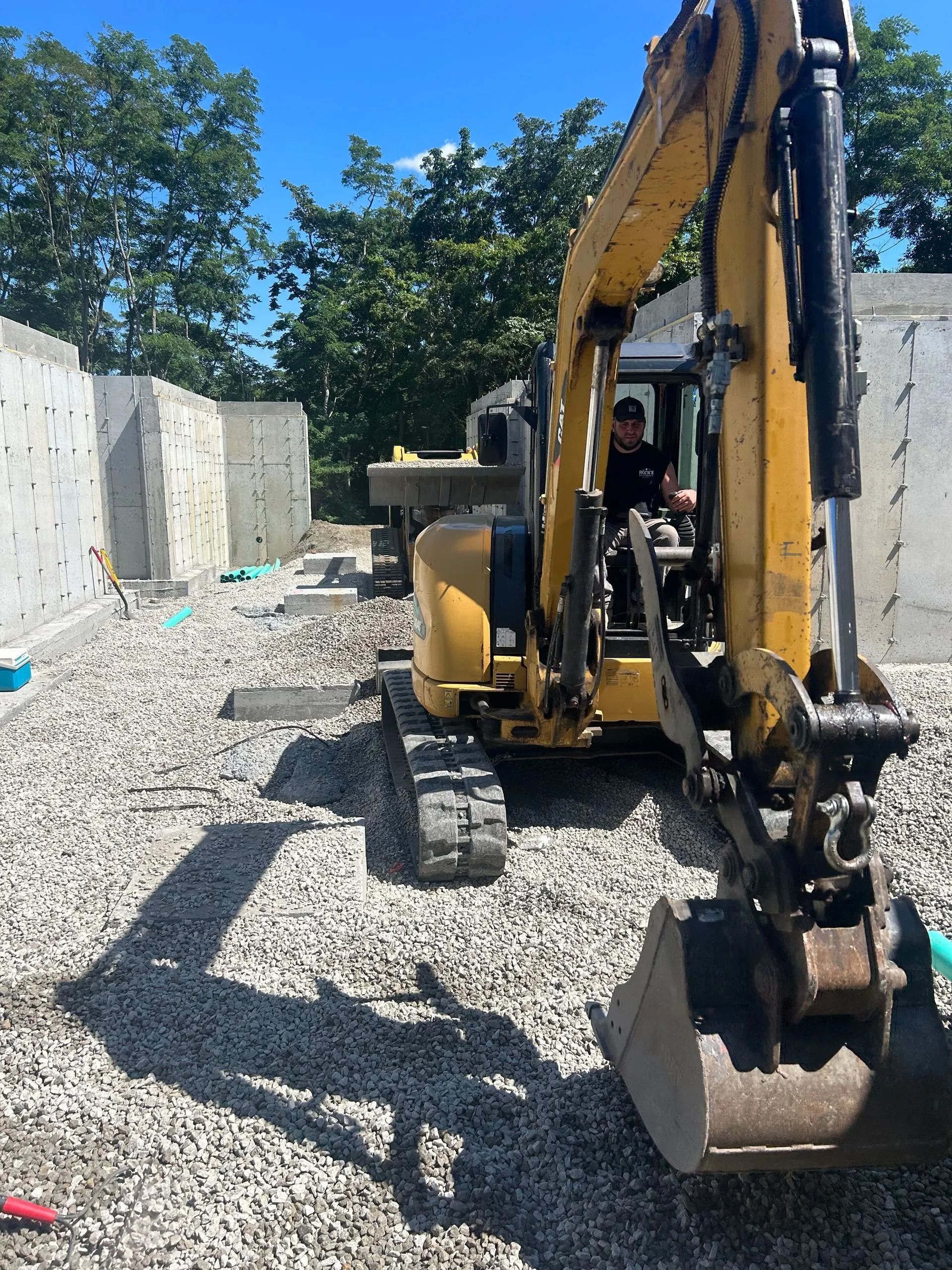 A yellow excavator sits on a gravel construction site between two concrete walls, with an operator inside the cab.
