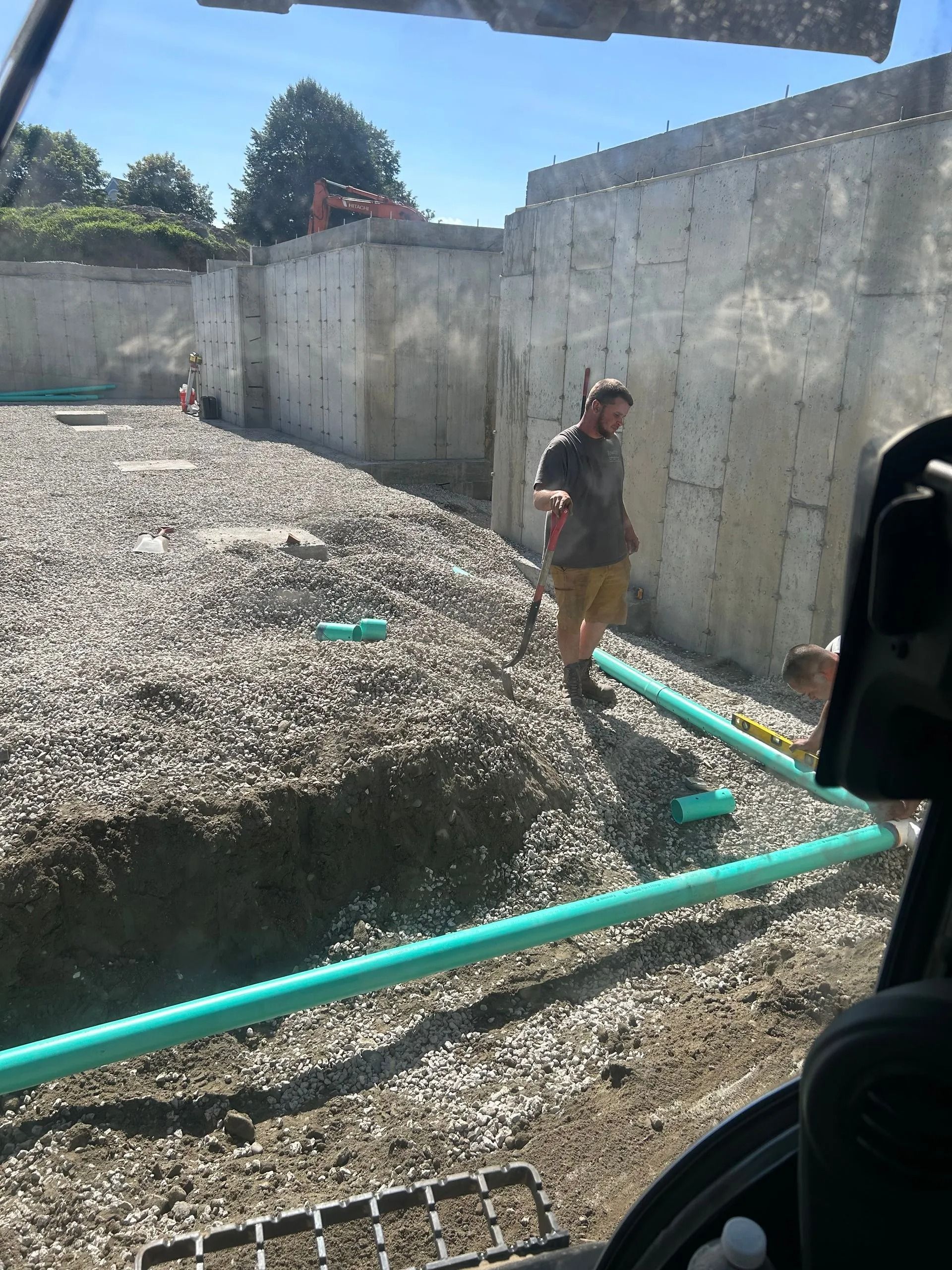 A construction worker stands in a foundation pit next to green PVC drainage pipes, surrounded by concrete walls.