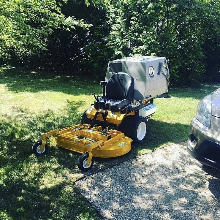 A yellow John Deere zero-turn lawn mower with a rear grass catcher sits on a grassy lawn next to a gravel driveway.