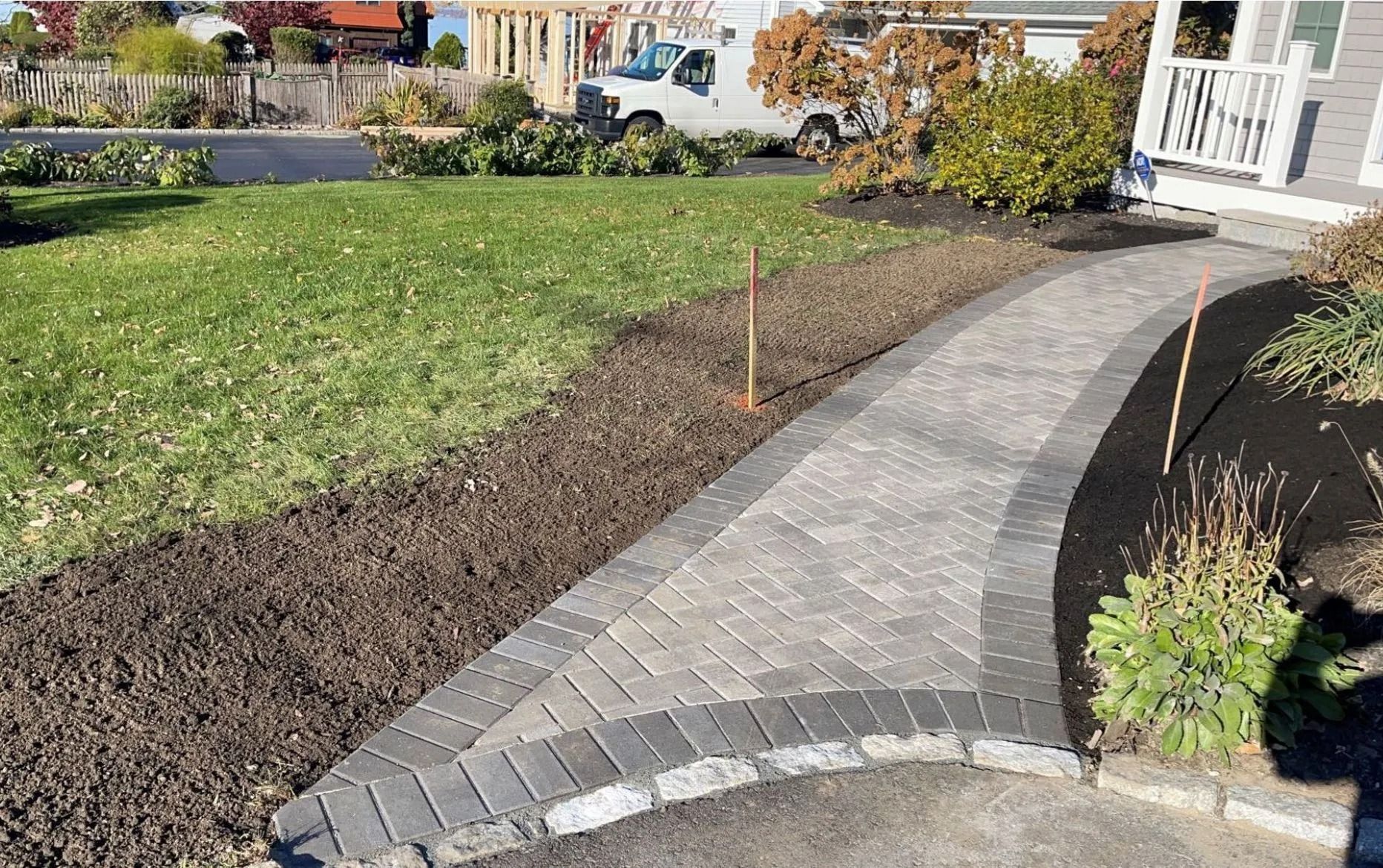 A paver walkway curves from a driveway through a yard with fresh mulch and grass toward a house entrance.