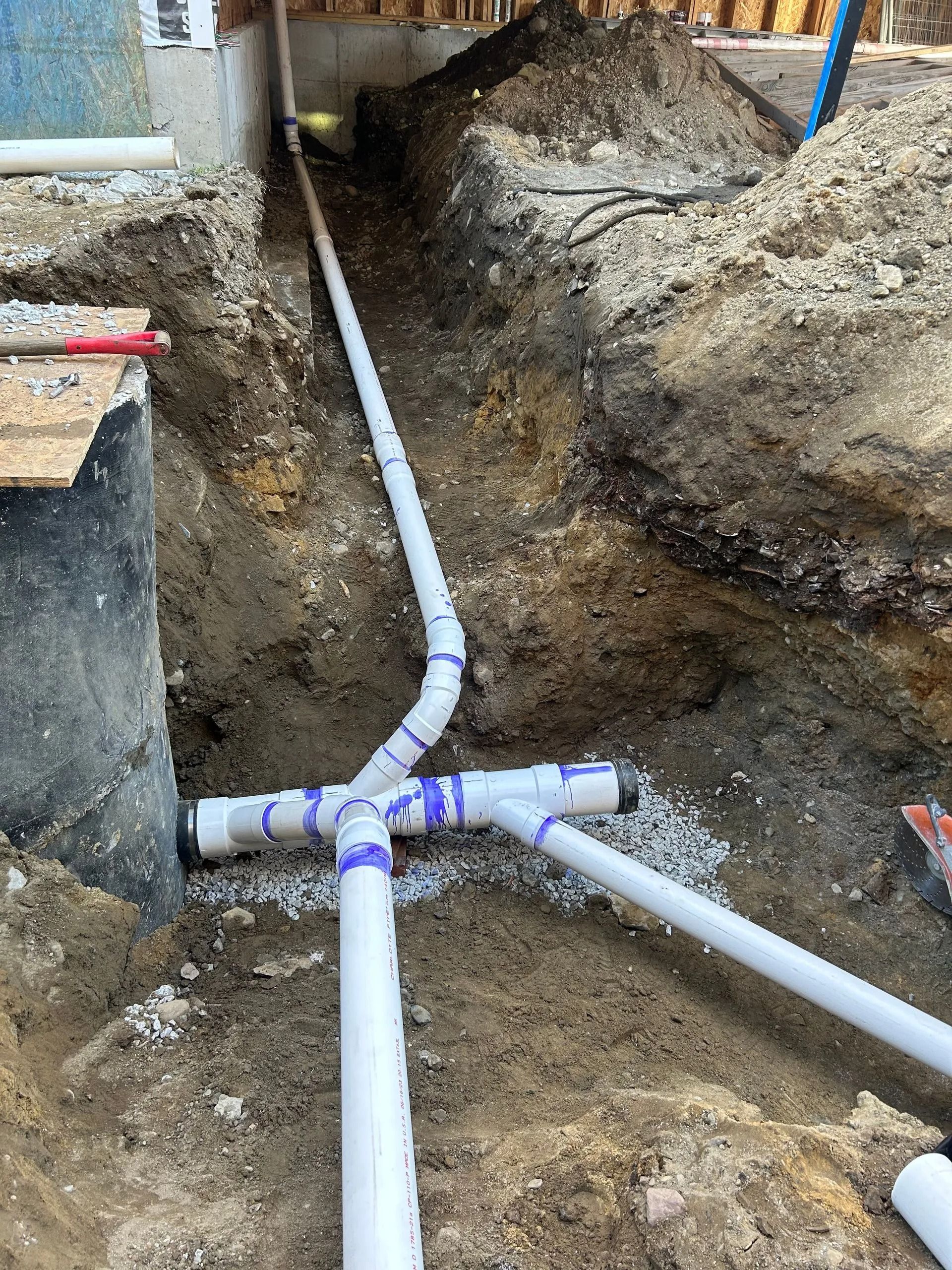 White PVC pipes connected in a T-junction and elbow configuration laid in an open dirt trench at a construction site.