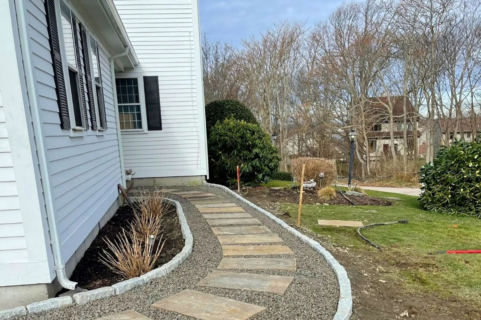 A stone paver walkway bordered by light grey stones and gravel follows the side of a white house with black shutters.