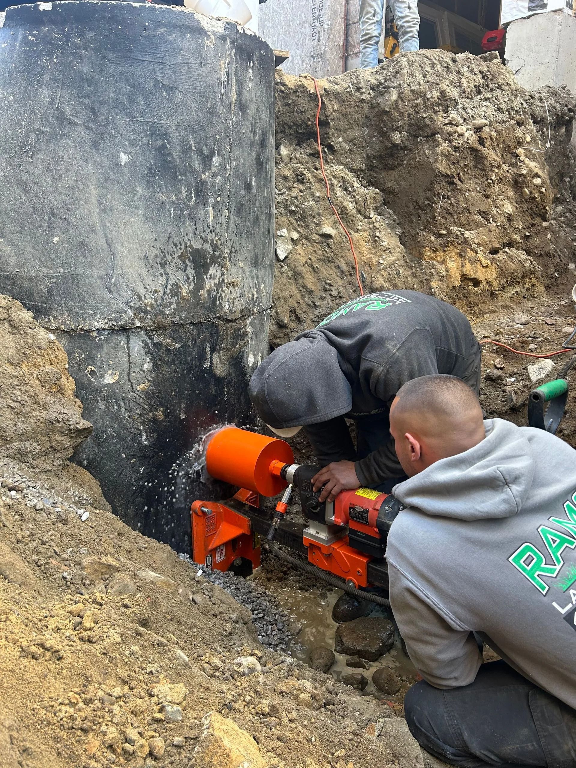 Two construction workers use a core drill to cut a hole into the side of a concrete utility structure in a dirt trench.