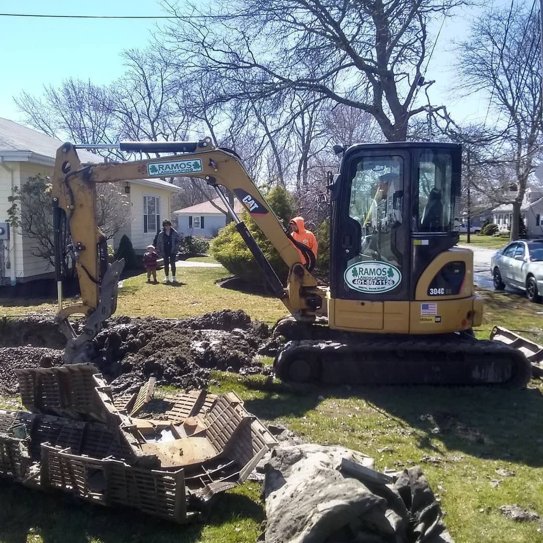 A yellow CAT excavator works on a residential lawn, with a person in an orange vest nearby and houses in the background.