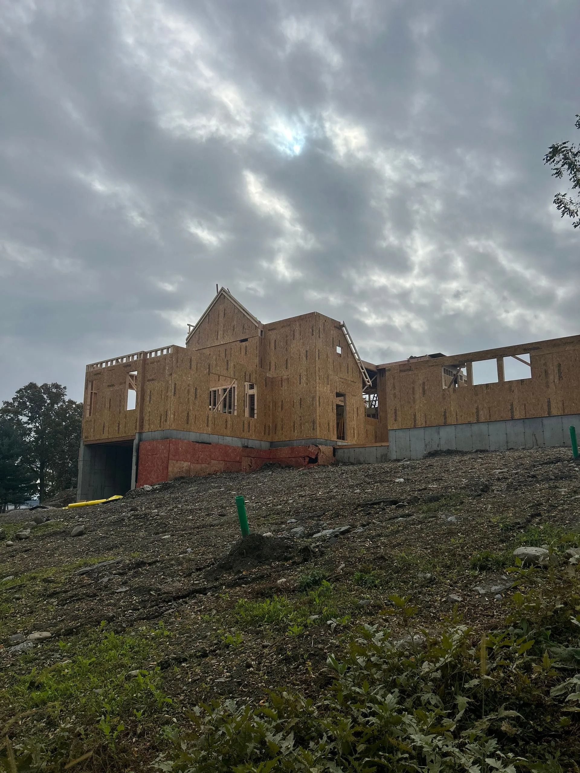 A two-story wood-framed house under construction stands on a sloped, rocky lot beneath a cloudy sky.
