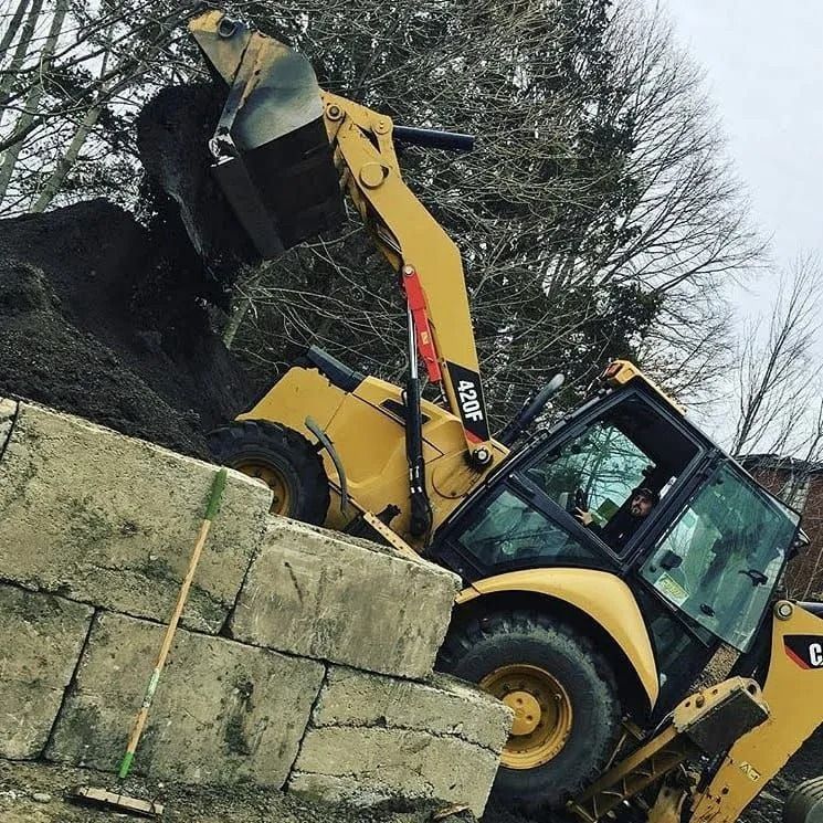 A yellow Caterpillar backhoe loader lifting a scoop of dark soil near large concrete block walls outdoors.