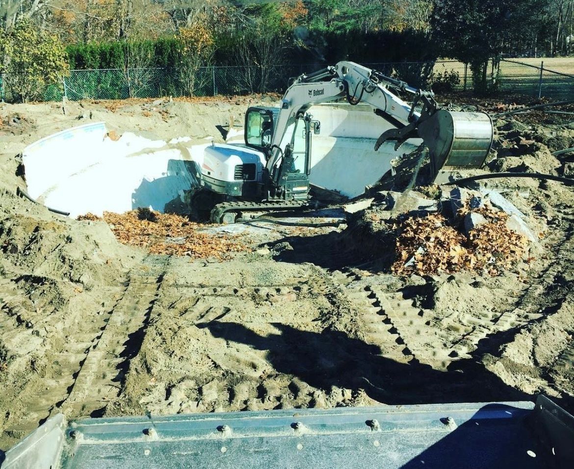 A white excavator digging in a dirt lot to install an inground swimming pool shell.