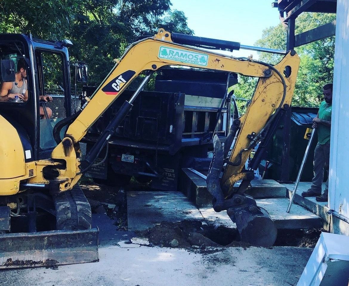 A yellow CAT excavator lifts a log near a parked dump truck while a person stands nearby on a construction site.