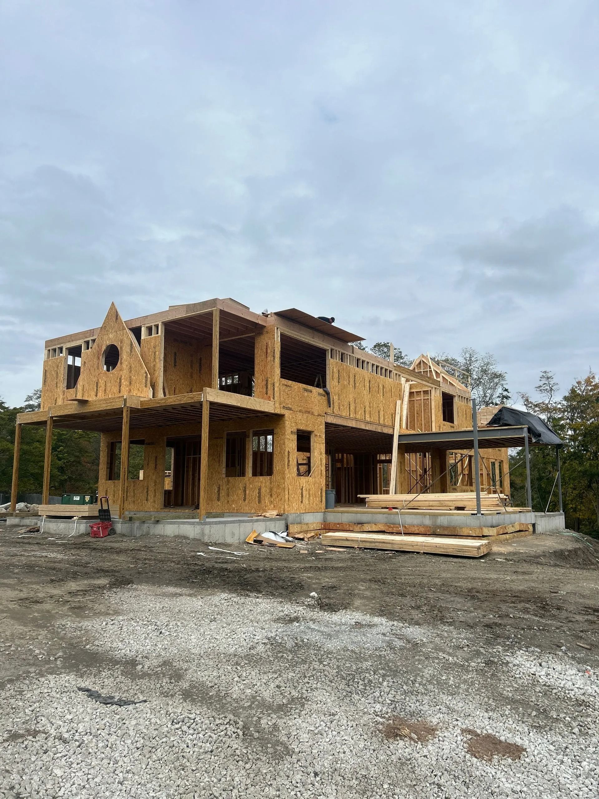 A two-story house under construction with exposed wooden framing and plywood sheathing on a gravel lot under a cloudy sky.