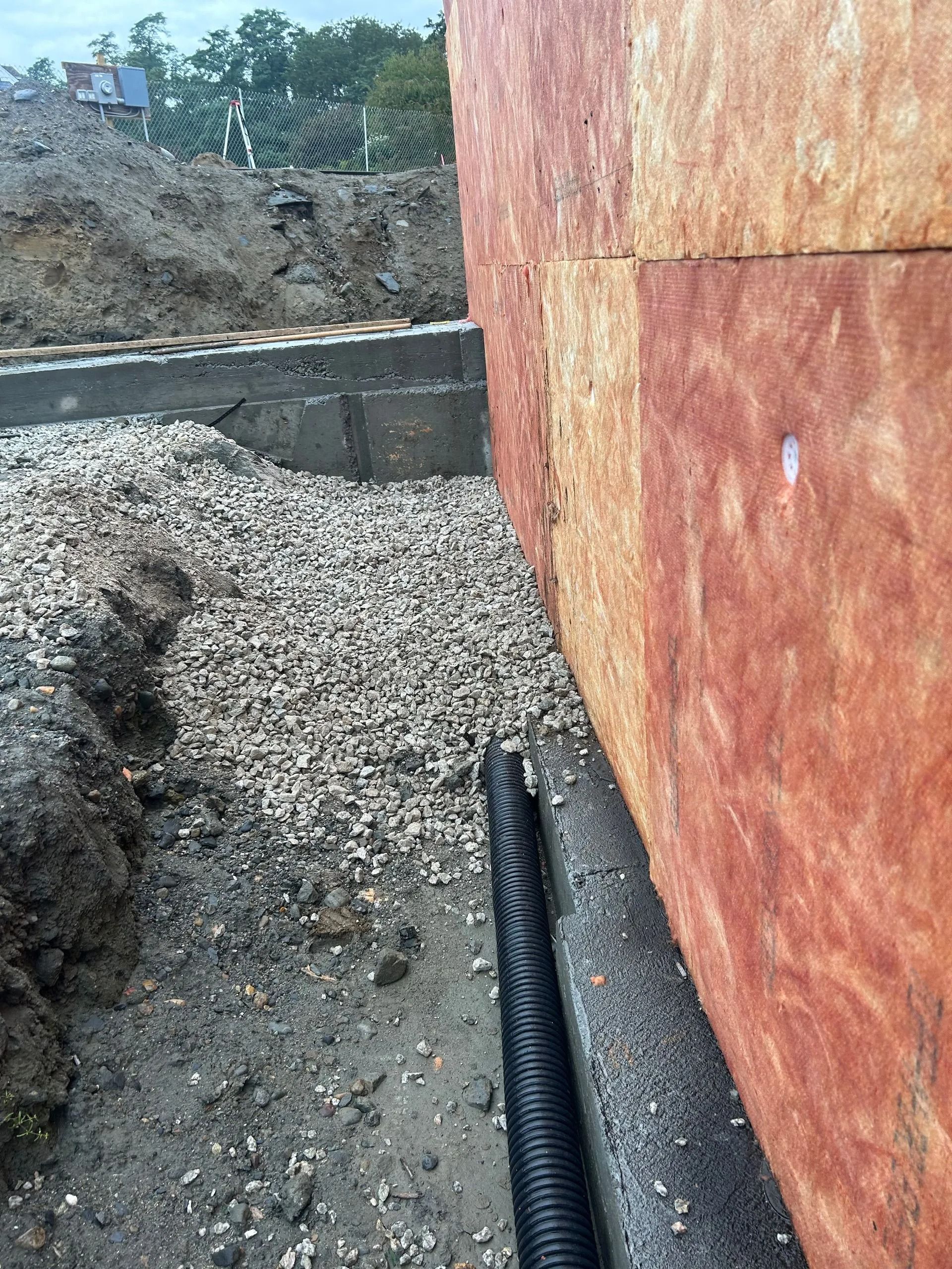 A foundation wall with insulation panels, a black drainage pipe, and a gravel base at a construction site.