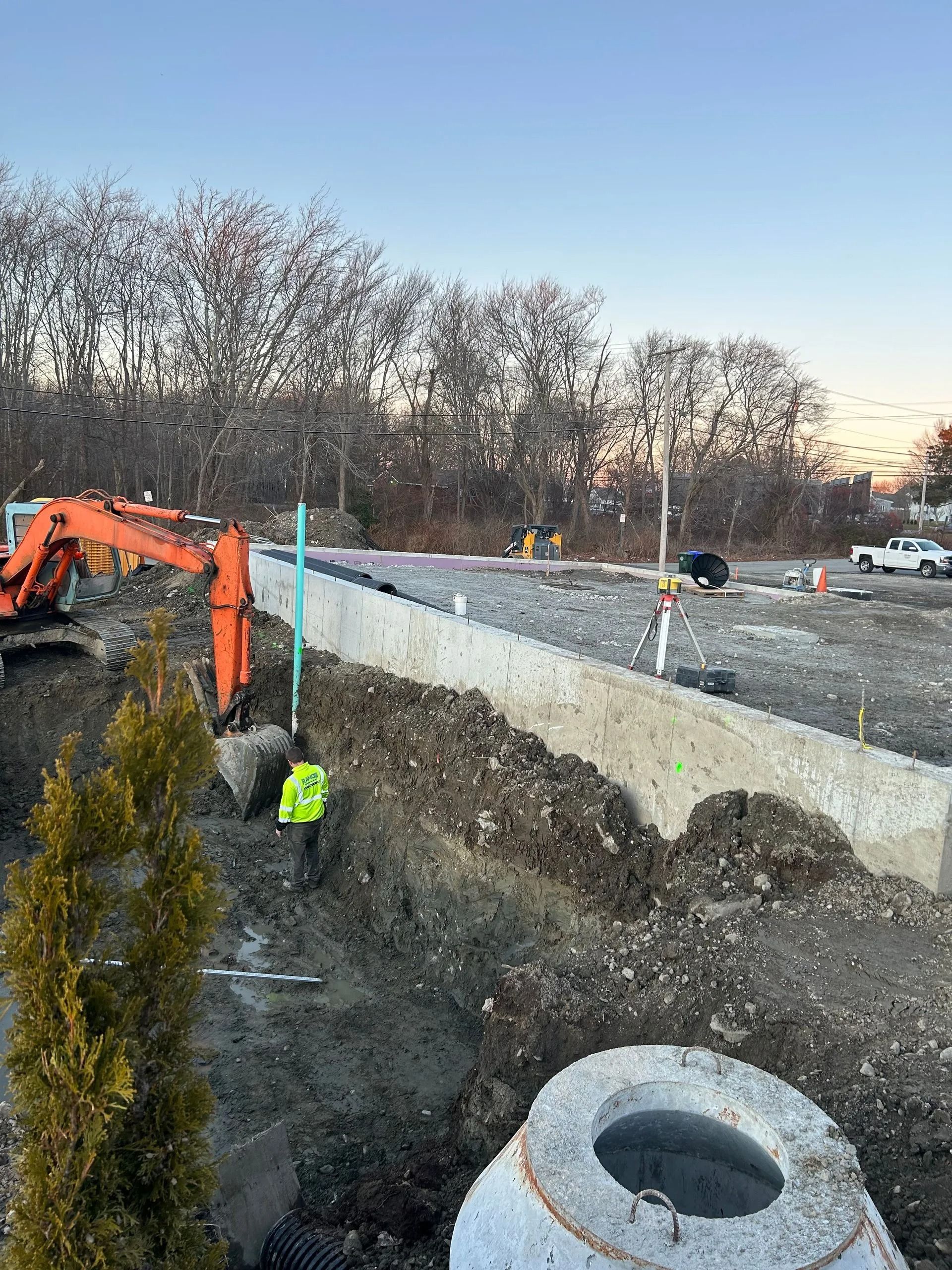 An orange excavator works at a construction site with a worker in a high-visibility yellow vest near a concrete wall.