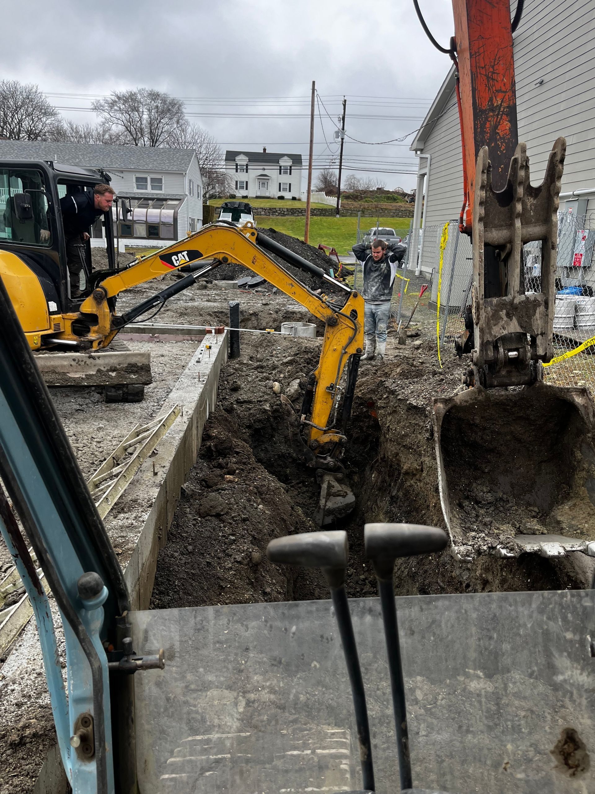 Two construction workers operate yellow excavators to dig a trench in a gravel lot next to a building.