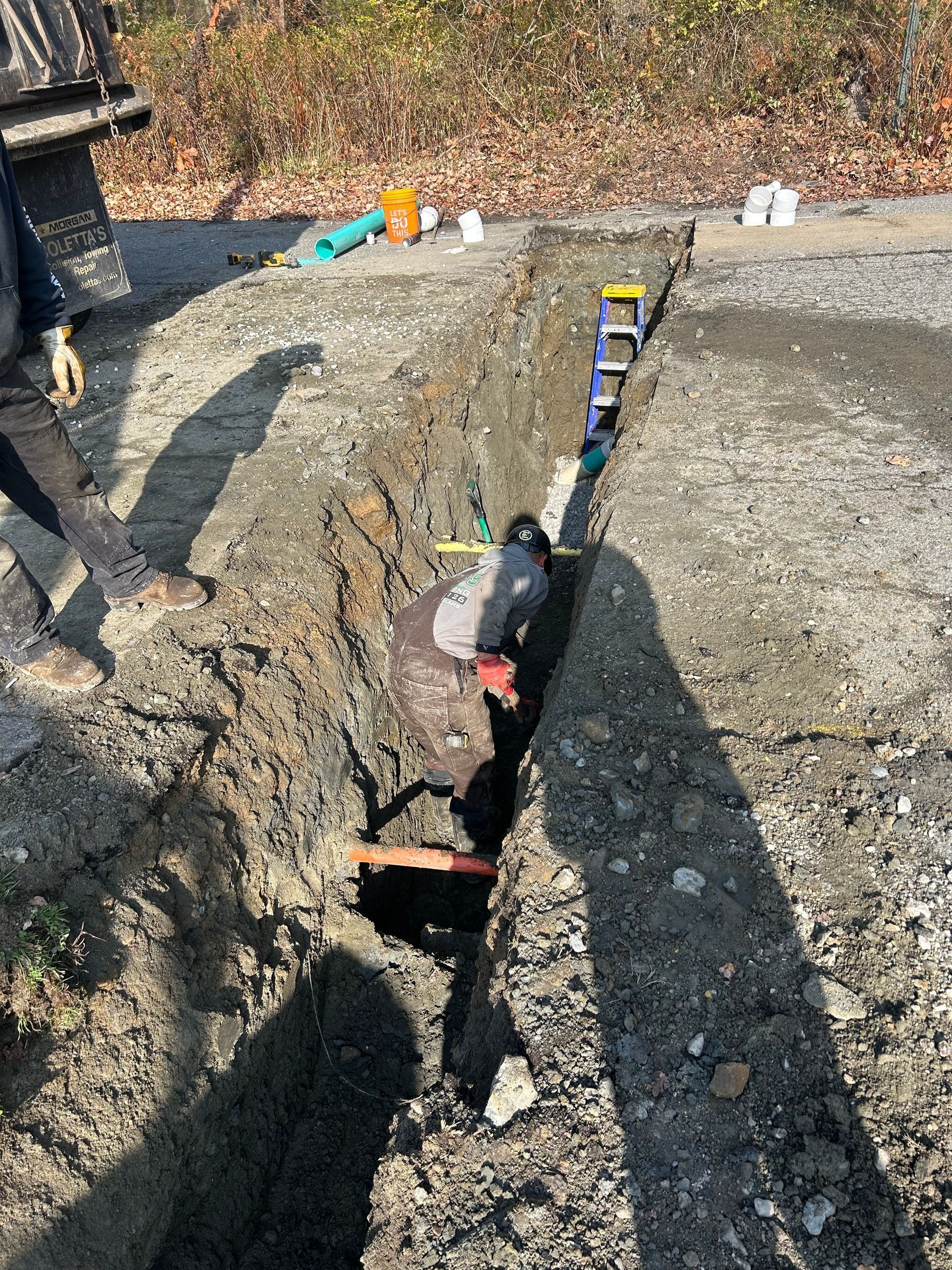 A person stands inside a narrow trench at an outdoor construction site, reaching toward a pipe laid in the ground.
