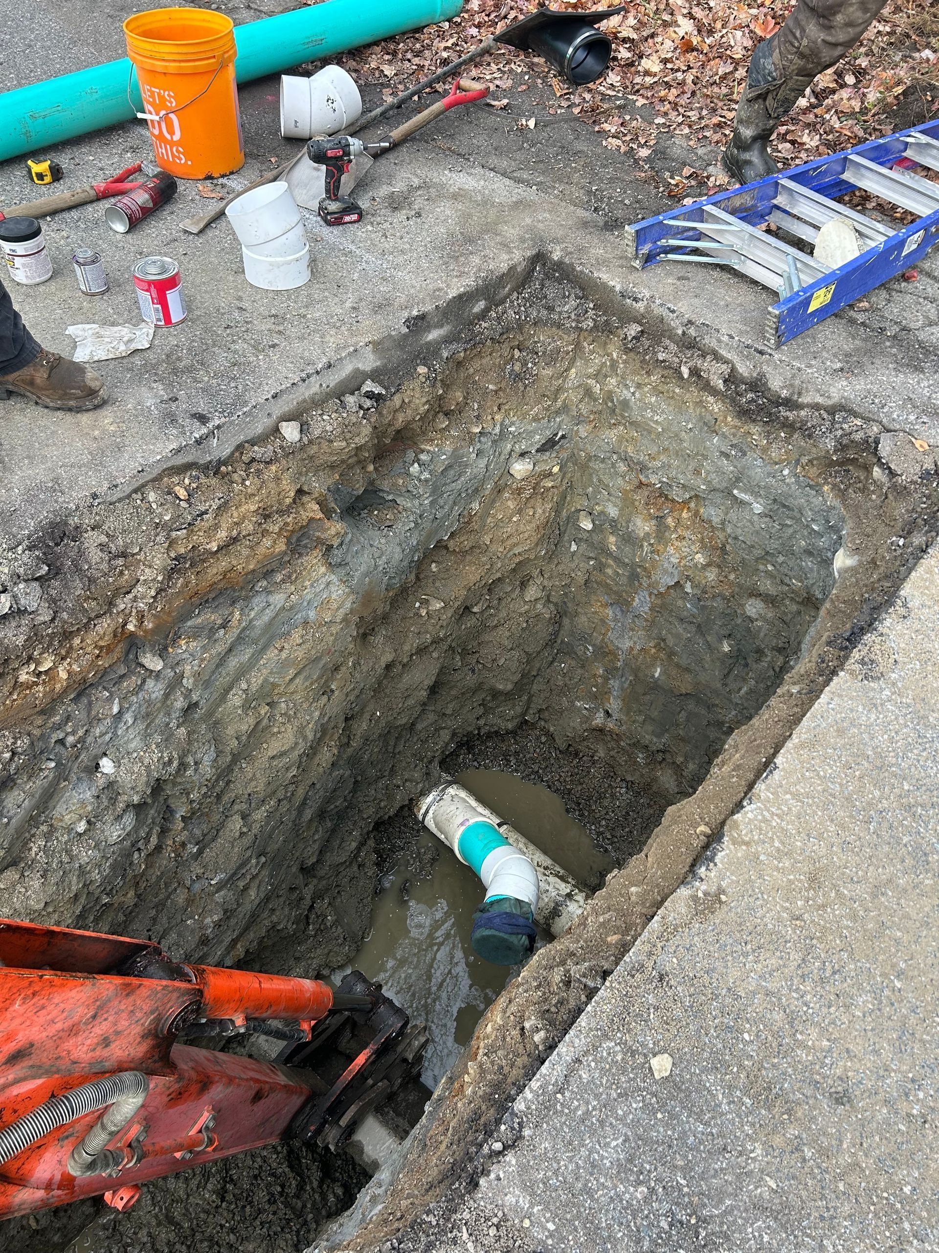 An excavator bucket works in a deep trench at a construction site, revealing an elbowed pipe connection in murky water.
