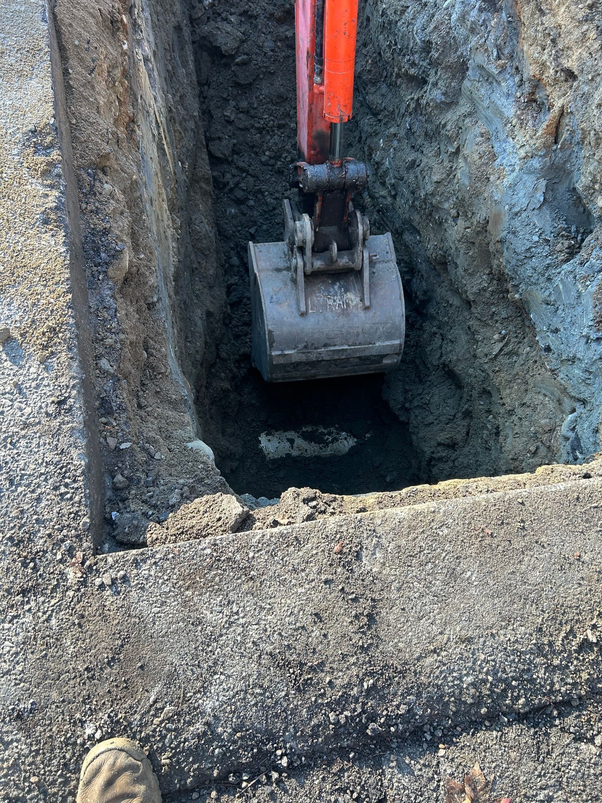 An excavator bucket digs into a narrow trench cut into a paved asphalt road surface.