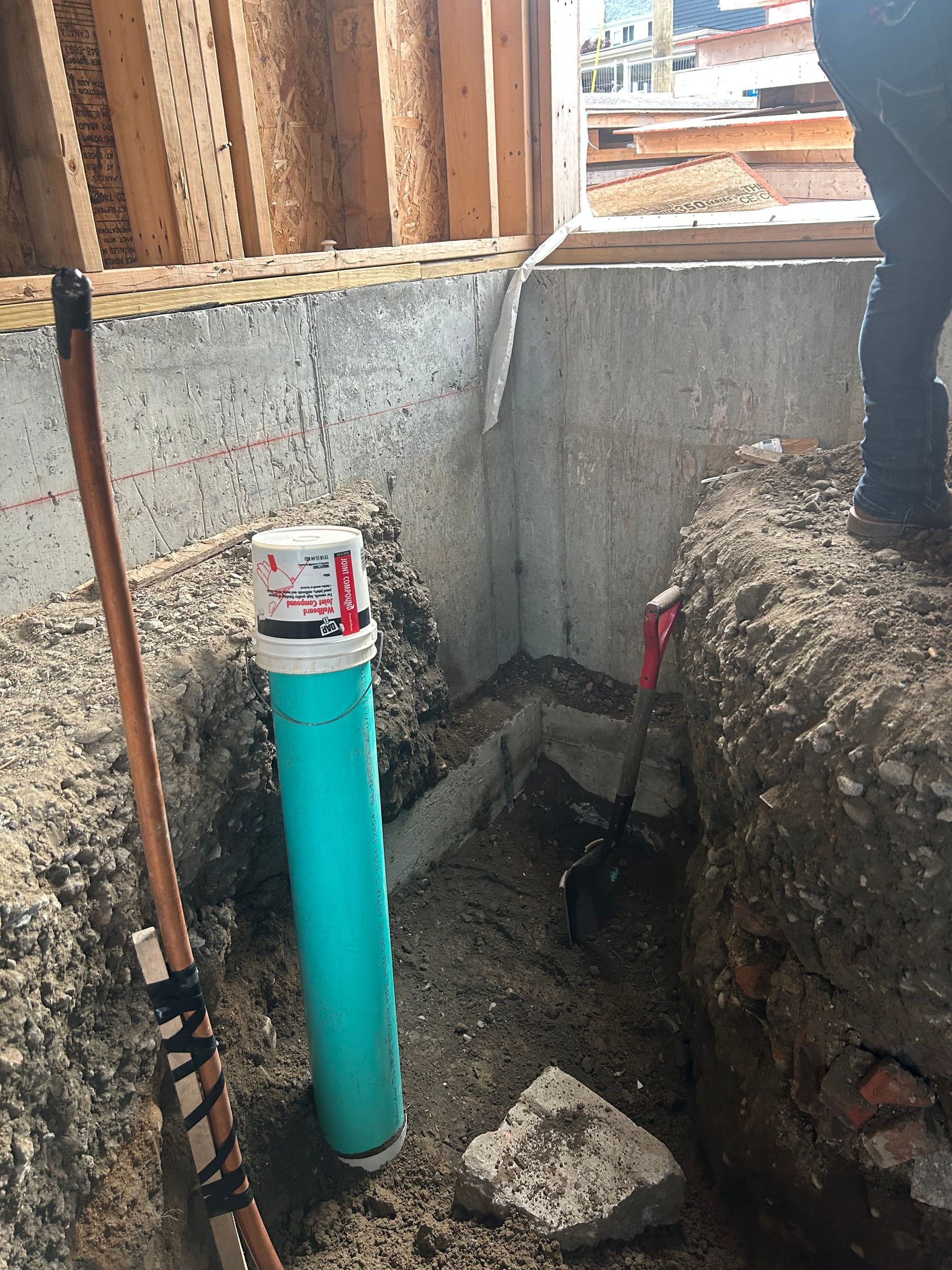 A construction site shows a vertical teal pipe in a dirt trench next to a concrete wall and a partially visible worker.
