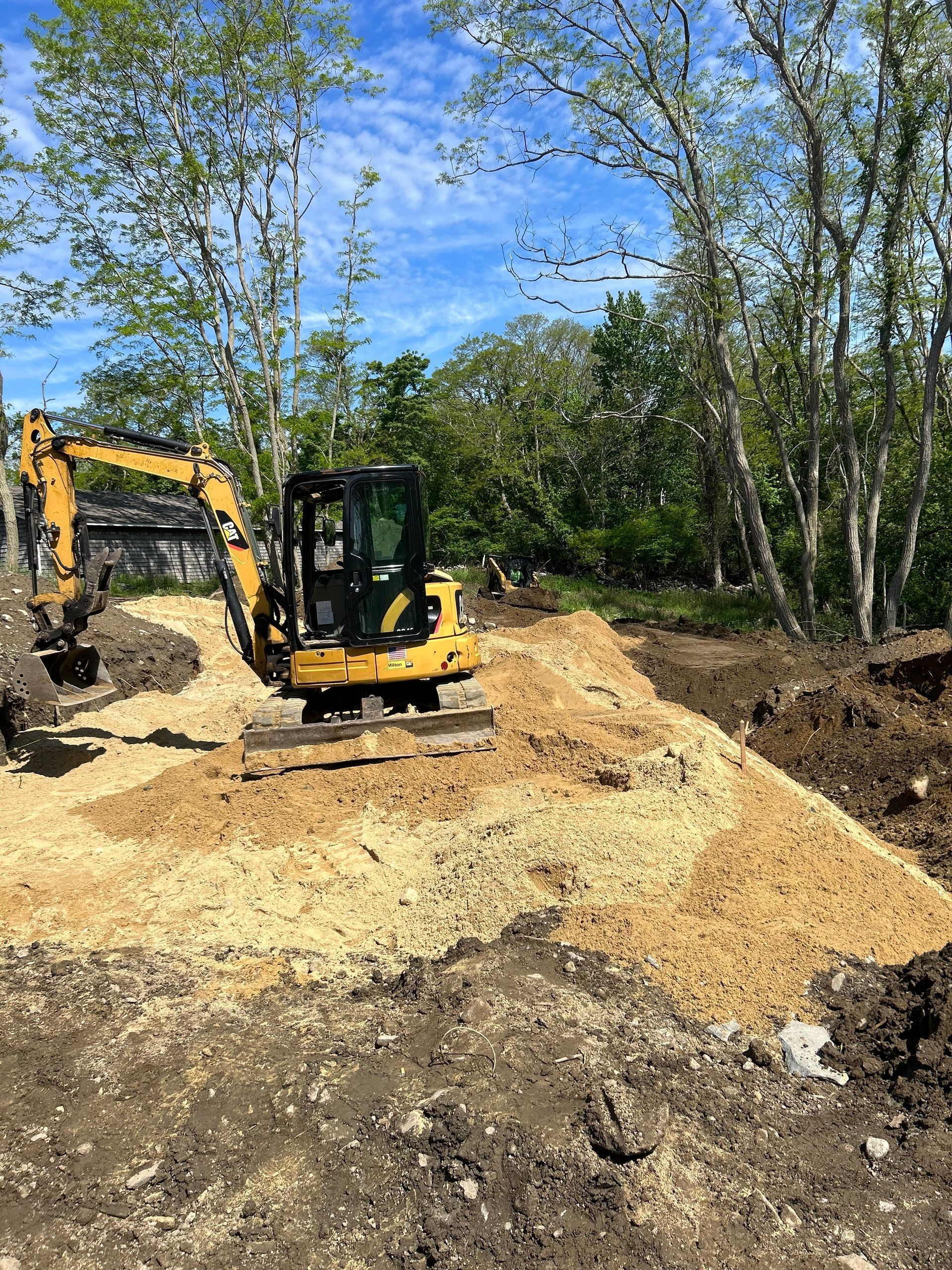 A yellow mini excavator sits on a mound of dirt at a sunny, tree-lined construction site.