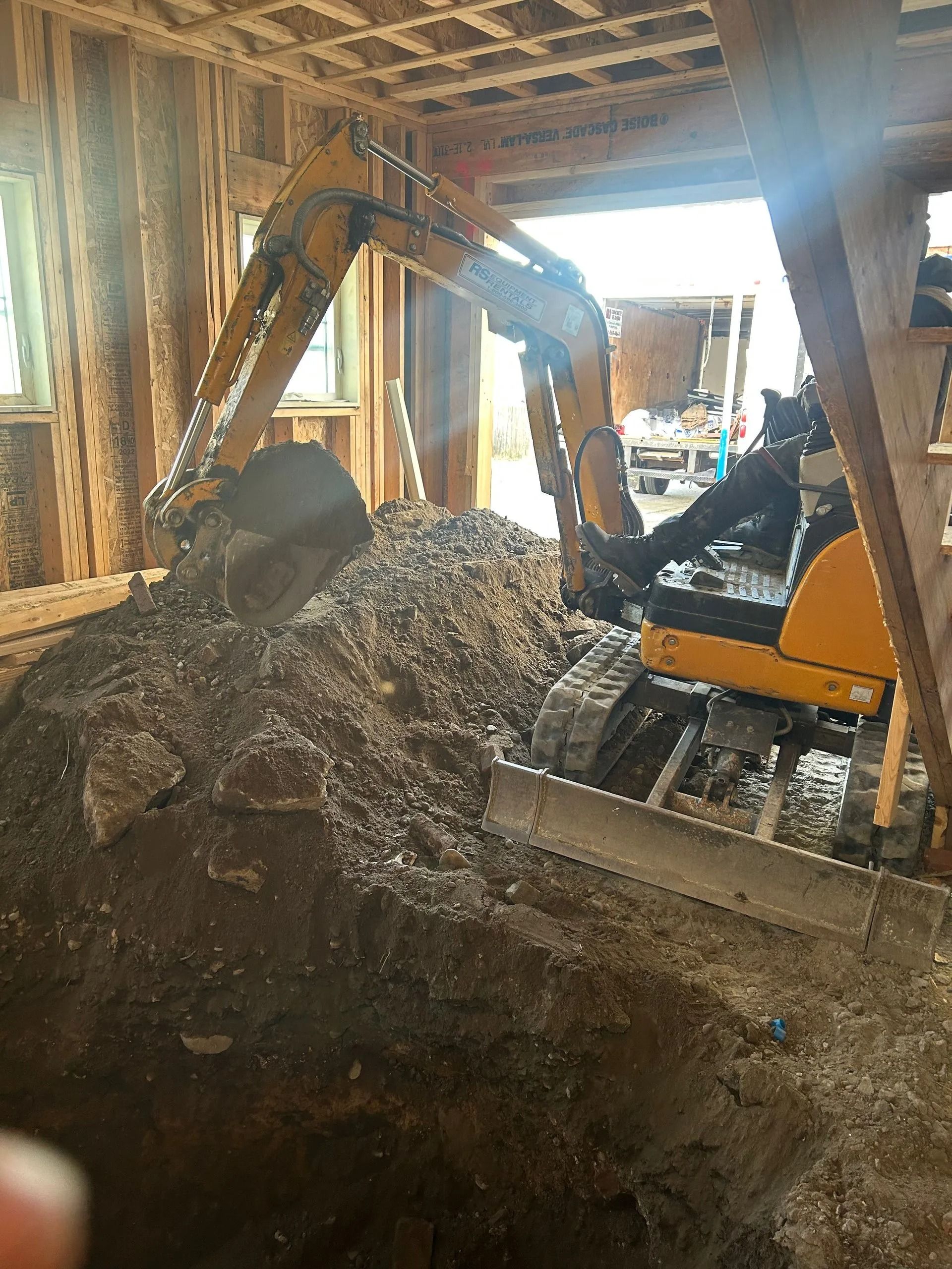 A yellow compact excavator is digging a trench inside a wooden framed building under construction.