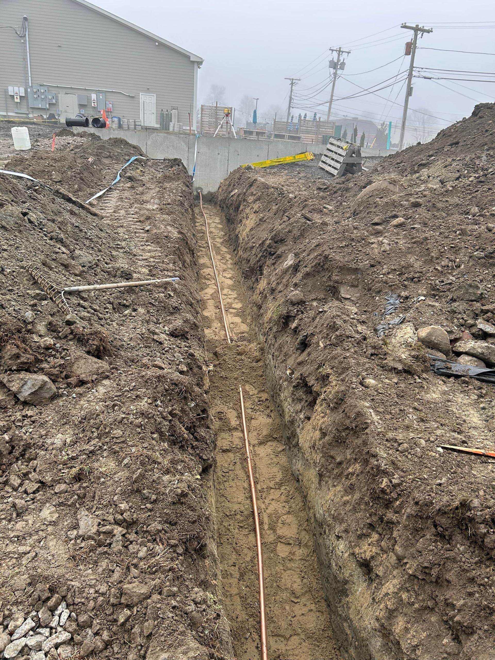 A narrow, muddy trench containing a copper grounding wire, located at a construction site with a building in the distance.