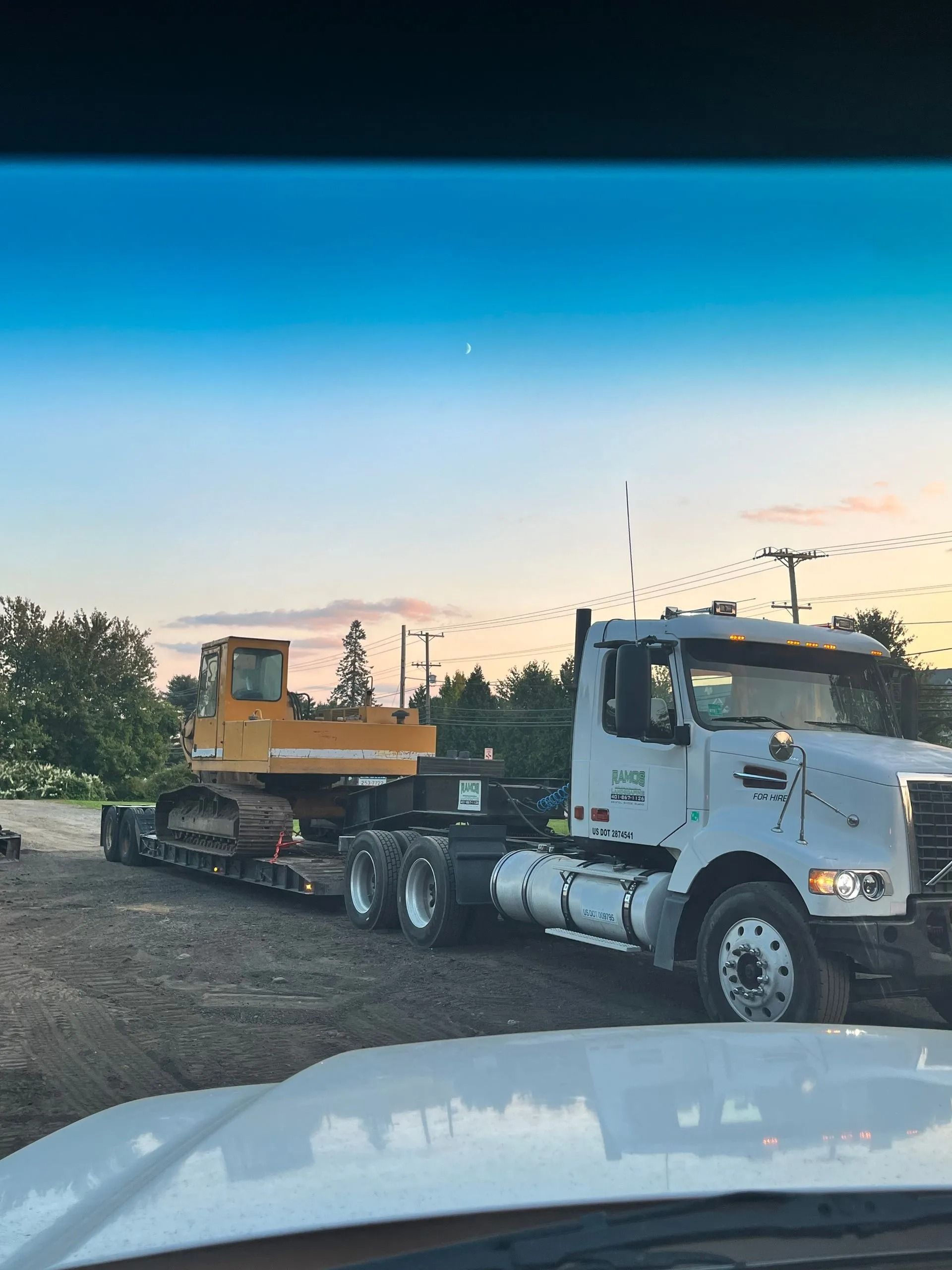 A white semi-truck hauling a yellow excavator on a flatbed trailer, parked on a dirt lot at dusk.